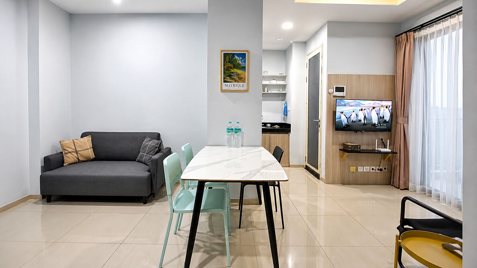 A modern living room with a sofa, a dining table with chairs, a television, and a kitchen area with a window.