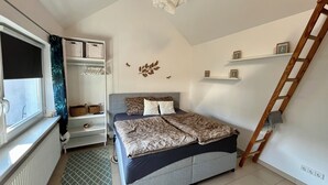 A bedroom with a bed, a window with a curtain, a radiator, a shelf with baskets, and a wall with decorative elements.