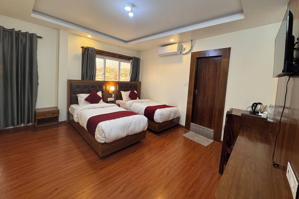A hotel room with two beds, a wooden floor, a window with curtains, an air conditioning unit, and a desk with a television.