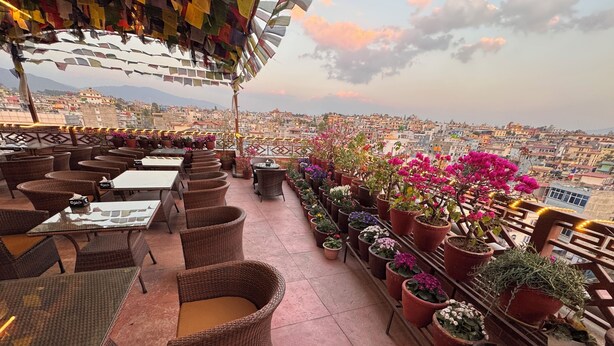An outdoor terrace with wicker furniture, potted plants, and a view of a cityscape.