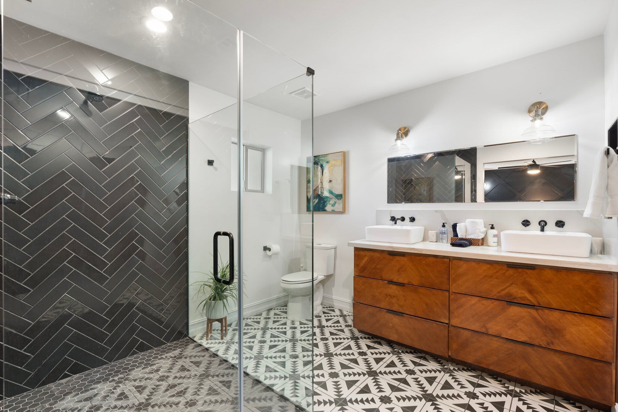 A modern bathroom with a glass shower enclosure, a white toilet, a wooden vanity with a mirror, and patterned floor tiles.