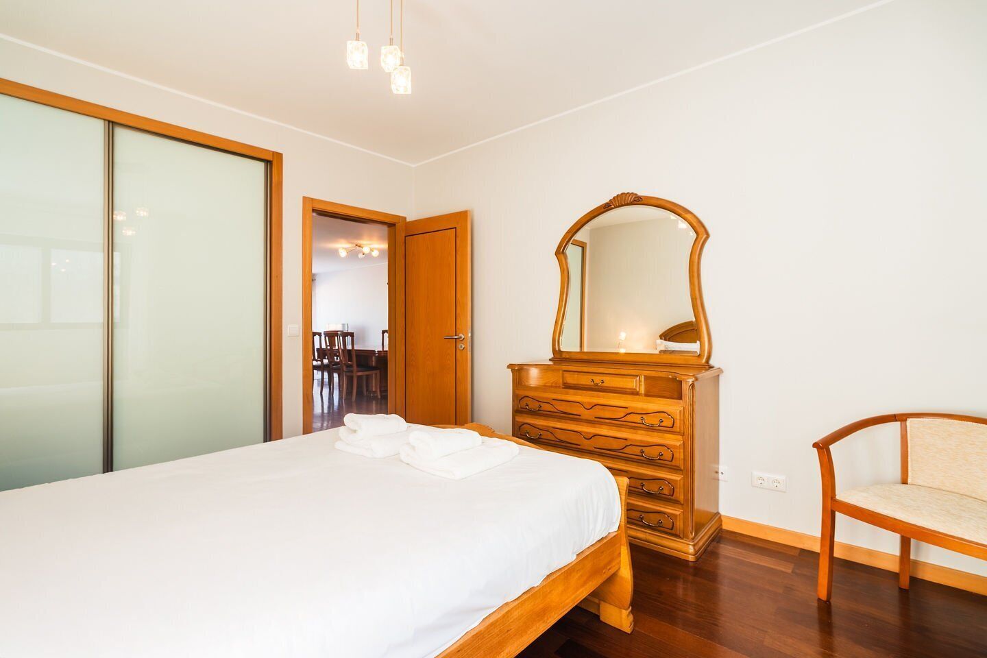 A bedroom with a bed, wooden dresser, mirror, chair, and a door leading to a dining area.