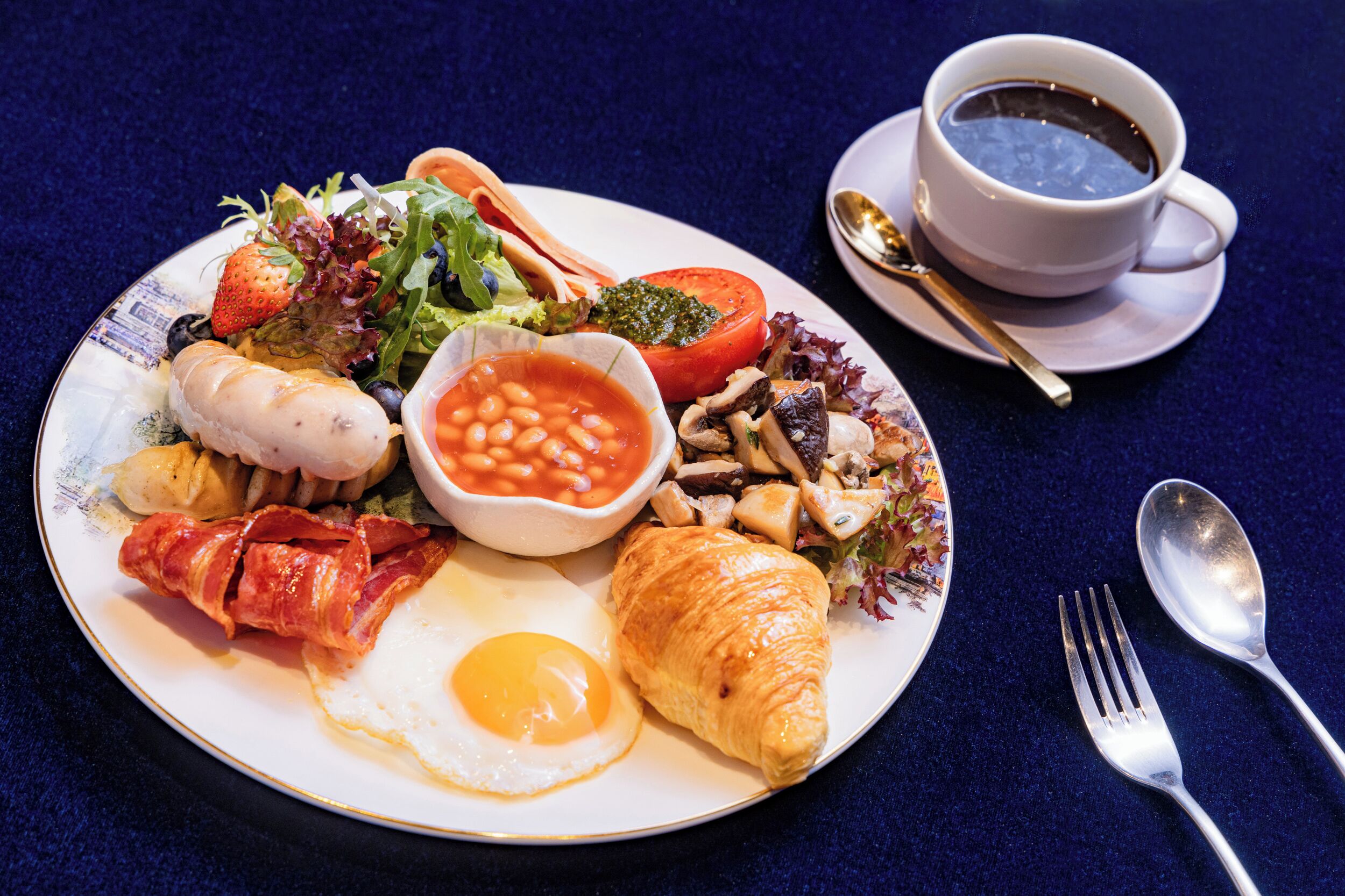 A plate of breakfast food including eggs, bacon, beans, a croissant, and a salad.