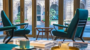 Een kamer met twee smaragdgroene fauteuils, een klein houten tafeltje en uitzicht op een historisch pand door grote ramen.