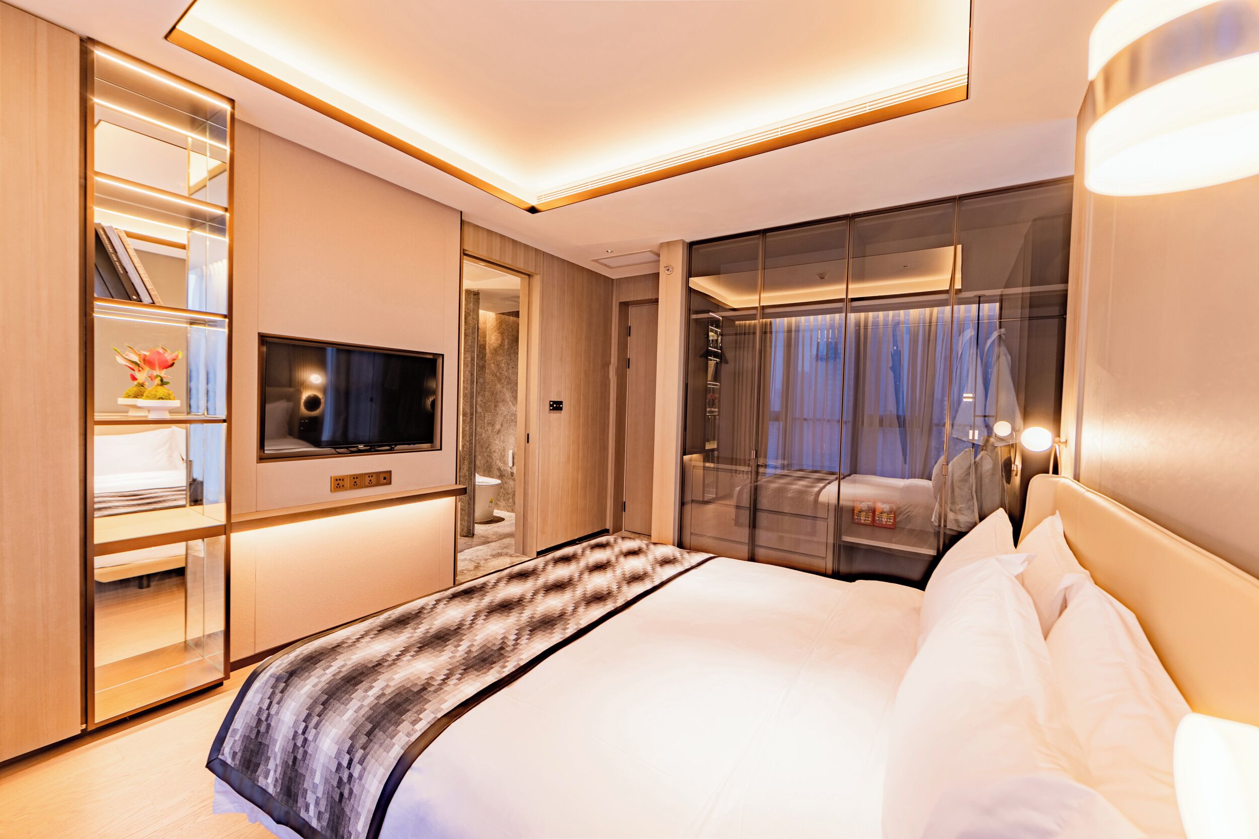 Une chambre d’hôtel moderne dotée d’un grand lit, d’une télévision, d’une armoire et d’une salle de bain visible à travers une paroi en verre.