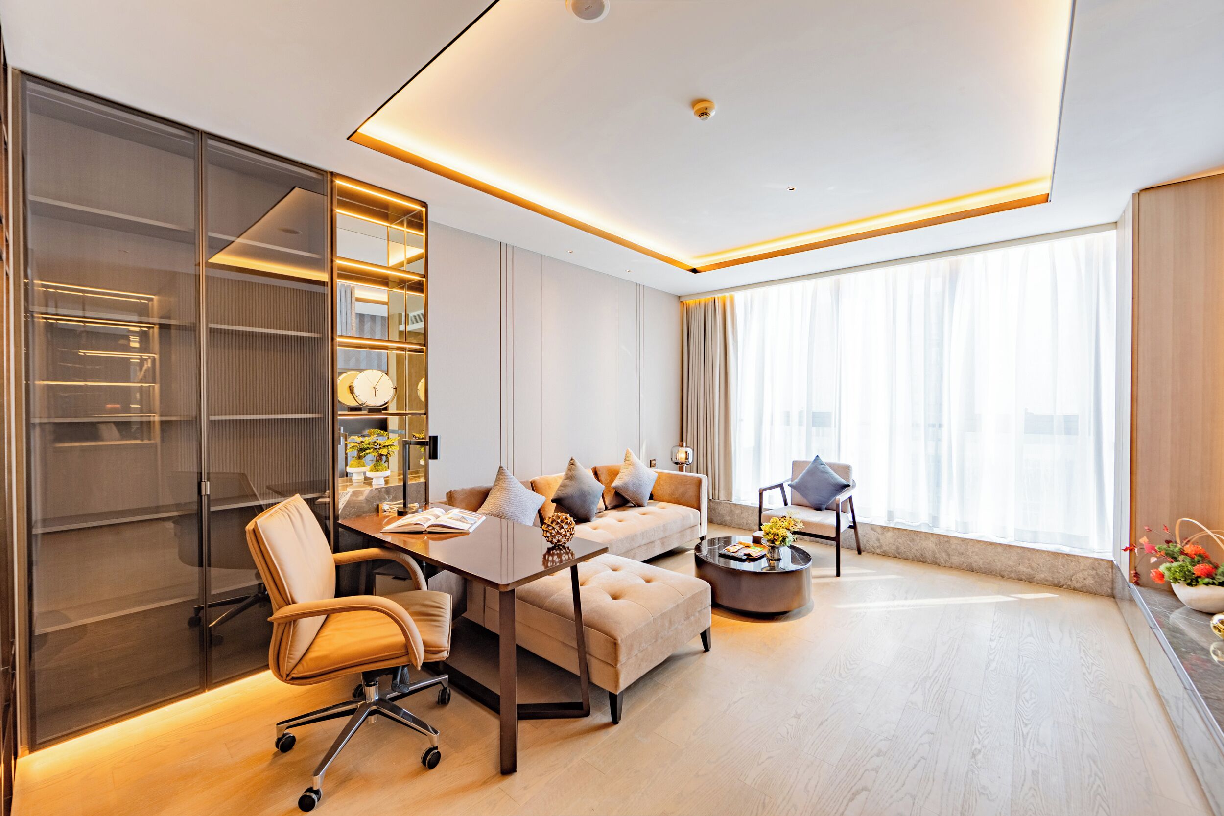 Ein modernes Wohnzimmer mit einer Couch, einem Couchtisch und einem Schreibtisch mit Stuhl. Ein Bücherregal und ein großes Fenster mit Vorhängen sind ebenfalls vorhanden.