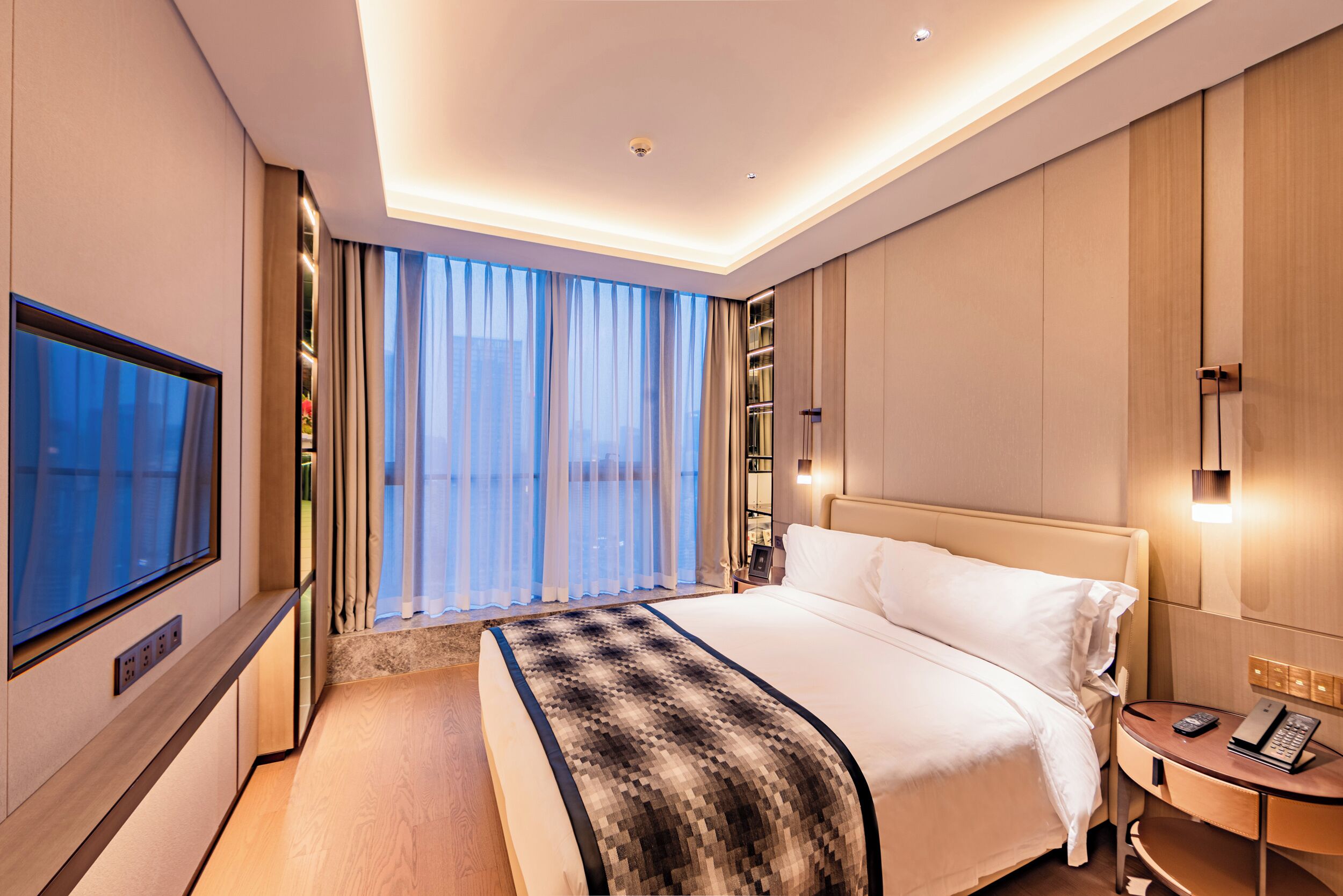 A modern hotel room with a large bed, a TV, a bedside table with a phone, and a view of the city through large windows.