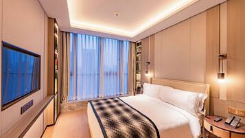 A modern hotel room with a large bed, a TV, a bedside table with a phone, and a view of the city through large windows.