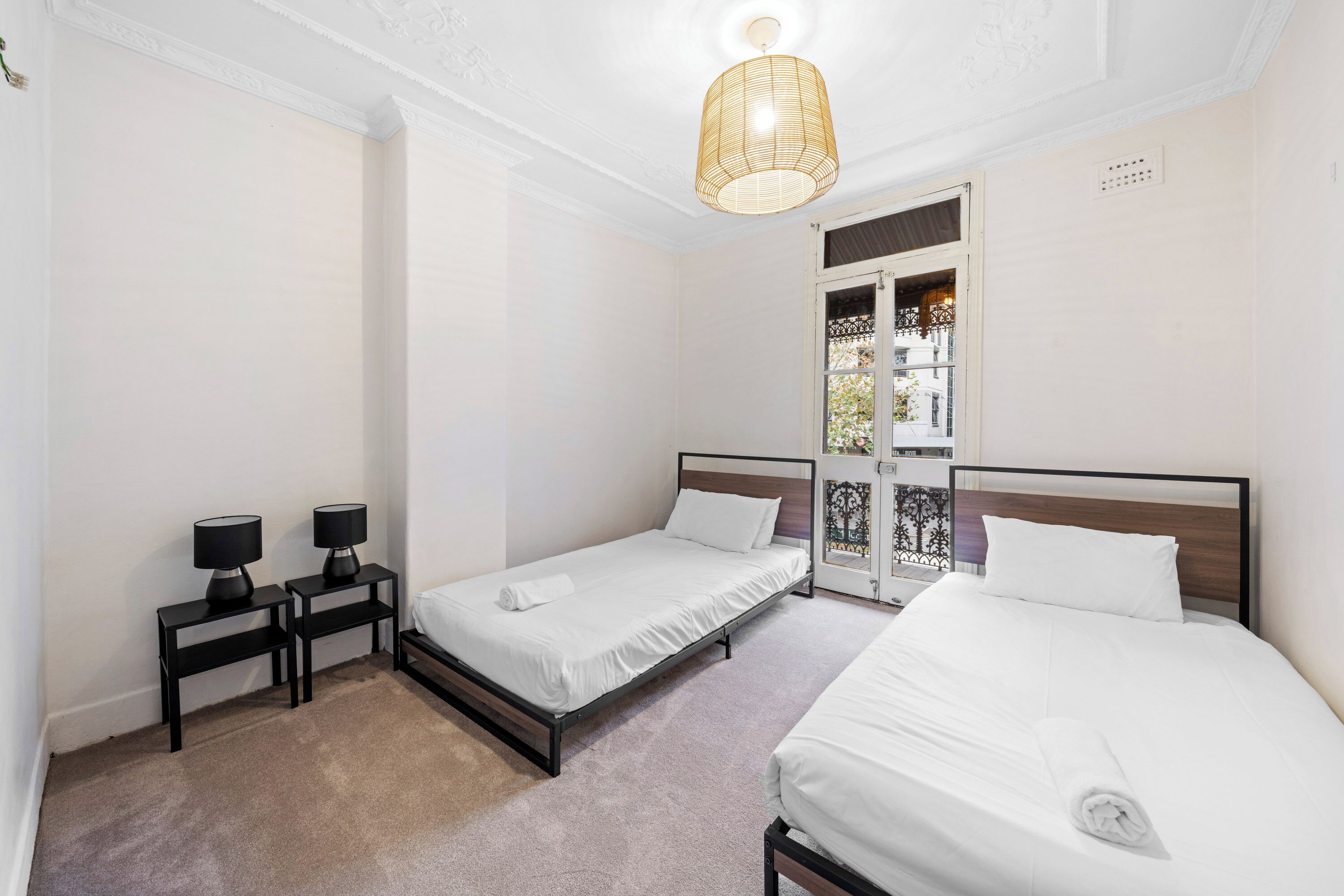 A hotel room with two single beds, a small table with two lamps, a door with a decorative glass panel, and a hanging light fixture.