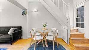 Ett modernt vardagsrum med ett matbord med glasskiva, omgivet av vita stolar, en mörkgrå soffa och en trappa som leder upp till ett andra våningsplan.