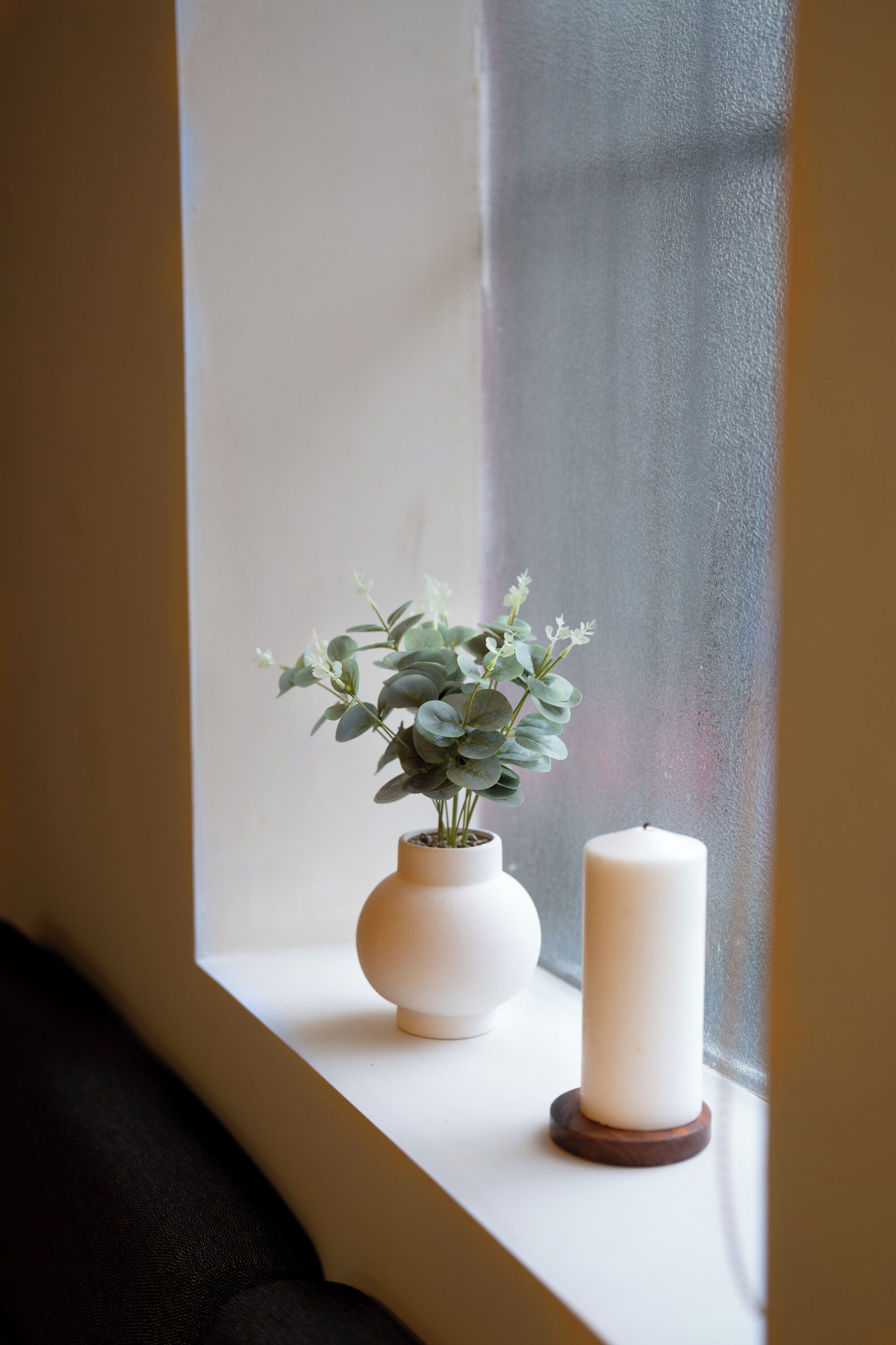 A window with a white vase containing a plant and a white candle on a wooden coaster.