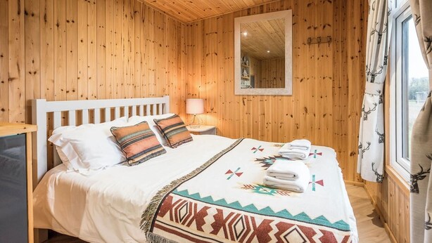 Habitación con paneles de madera, una cama, una mesita de noche, un espejo y un ventanal con cortinas.