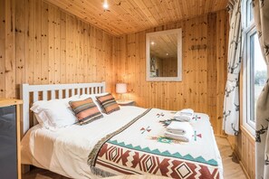 A wooden-paneled room with a bed, a nightstand, a mirror, and a window with curtains. - River Lodge, Aldeburgh (Aldeburgh)