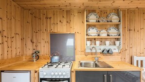 Una cocina con paredes de madera, estufa a gas, horno, lavamanos y estanterías con platos y tazas.