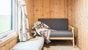 Una habitación de cabaña acogedora, con interior de madera, un sofá gris, una mesita con una botella de vino y una copa, y una ventana con cortinas.