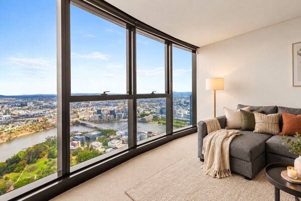 A modern living room with a large window offering a city view, a grey sofa with cushions, a floor lamp, and a small table with a candle.