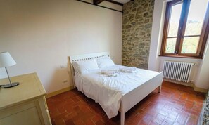 Une chambre avec un lit blanc, un mur en pierre, une fenêtre et un radiateur.