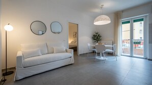 A modern living room with a white sofa, a dining table with chairs, and a large window with a view of buildings. - Appartamento con balcone vicino a Castelvecchio (Verona)