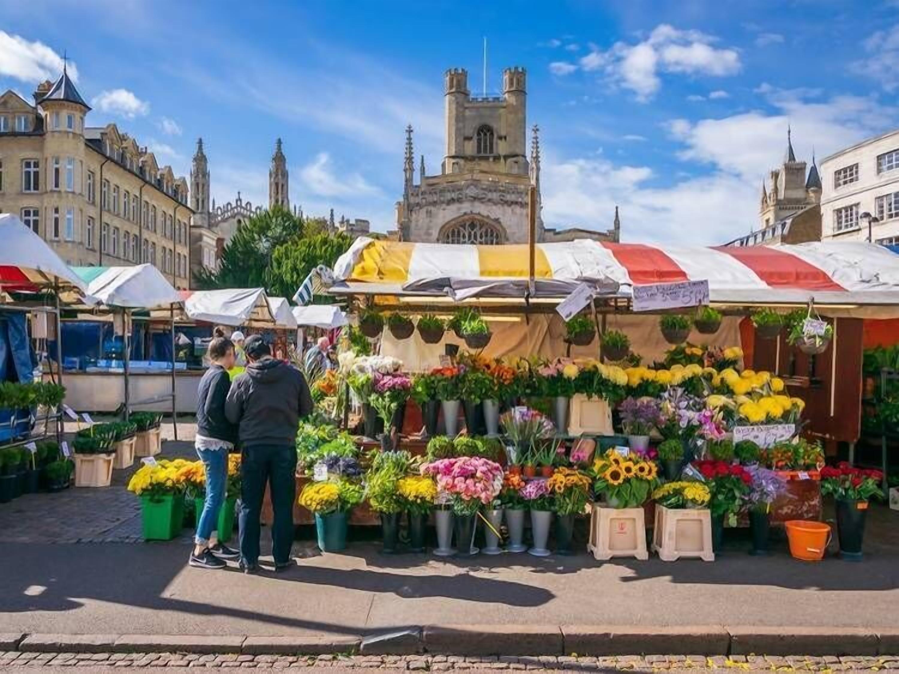 A flower market with various stalls selling flowers and plants, set against a backdrop of historic buildings and a prominent tower.