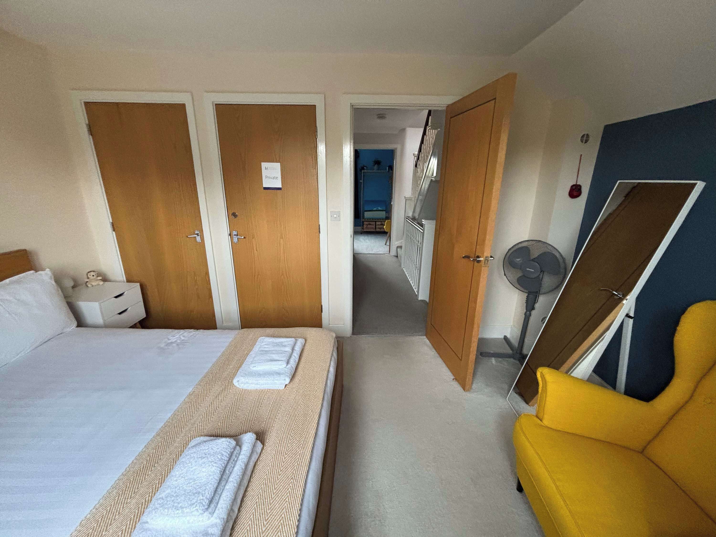 A hotel room with a bed, a yellow armchair, a desk with a lamp, and a mirror.