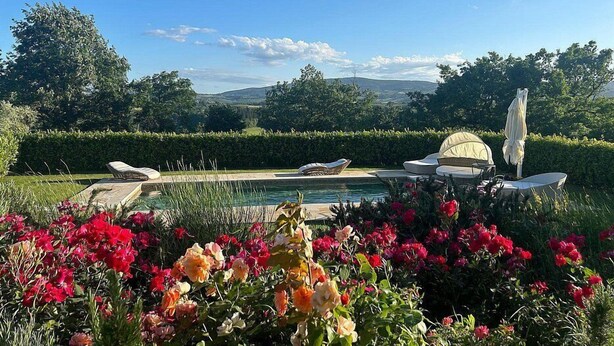 A pool area with lounge chairs and a table, surrounded by colorful flowers and a view of hills.