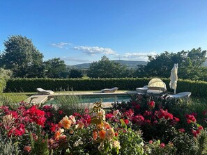 A pool area with lounge chairs and a table, surrounded by colorful flowers and a view of hills.