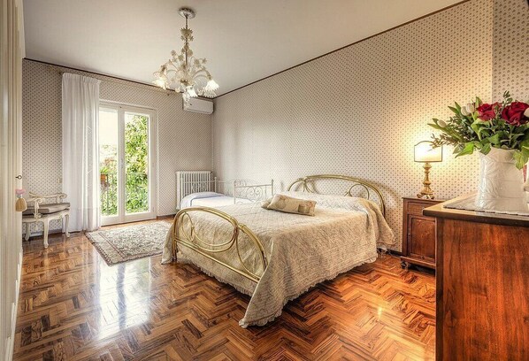 A bedroom with a large bed, a chandelier, a wooden floor, and a view through a glass door.