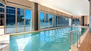 An indoor swimming pool with clear glass walls, a tiled floor, and metal pool ladders. - Modern 2-Bed with Pool Access in Surfers Paradise (Surfers Paradise)