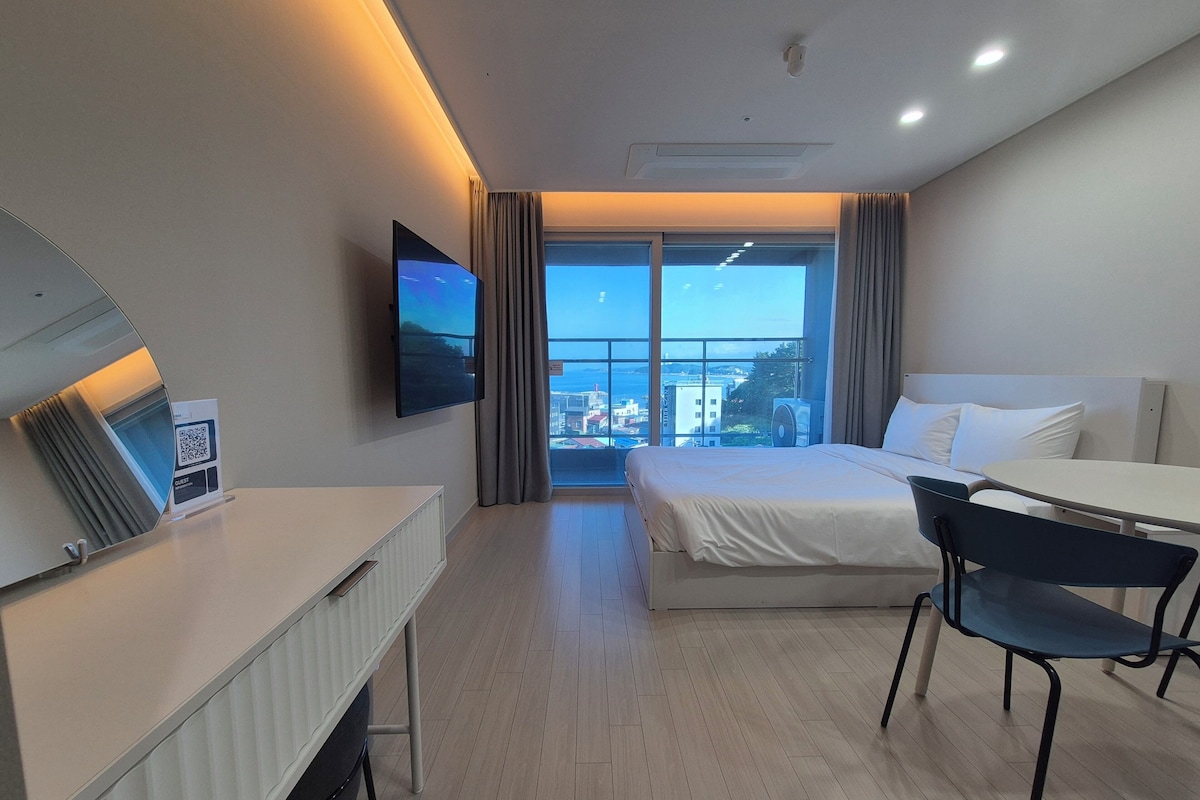 A hotel room with a bed, a desk, a chair, a TV, and a window with a city view.