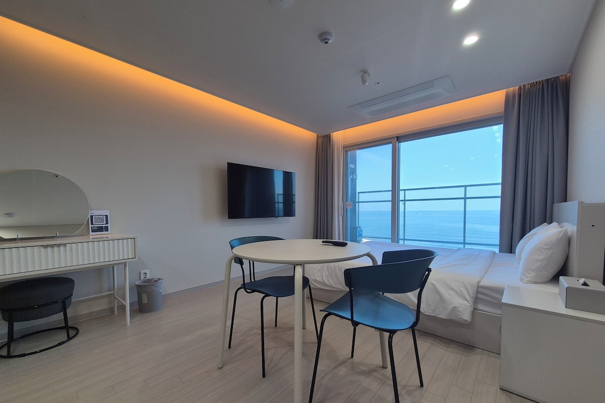 A modern hotel room with a balcony, a flat-screen TV, a round dining table with chairs, and a bed with a nightstand.