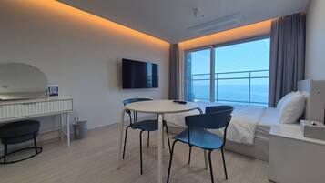 A modern hotel room with a balcony, a flat-screen TV, a round dining table with chairs, and a bed with a nightstand.