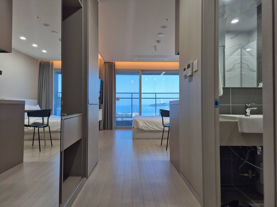 Habitación de hotel con cama, escritorio, silla, baño con lavamanos y espejo, y ventana grande con vistas.