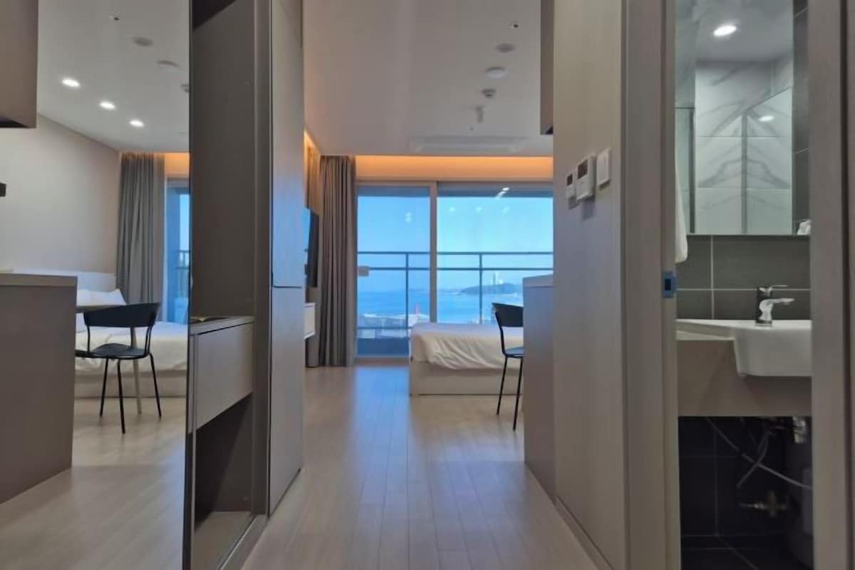 A hotel room with a bed, a desk, a chair, a bathroom with a sink, and a view of the ocean.