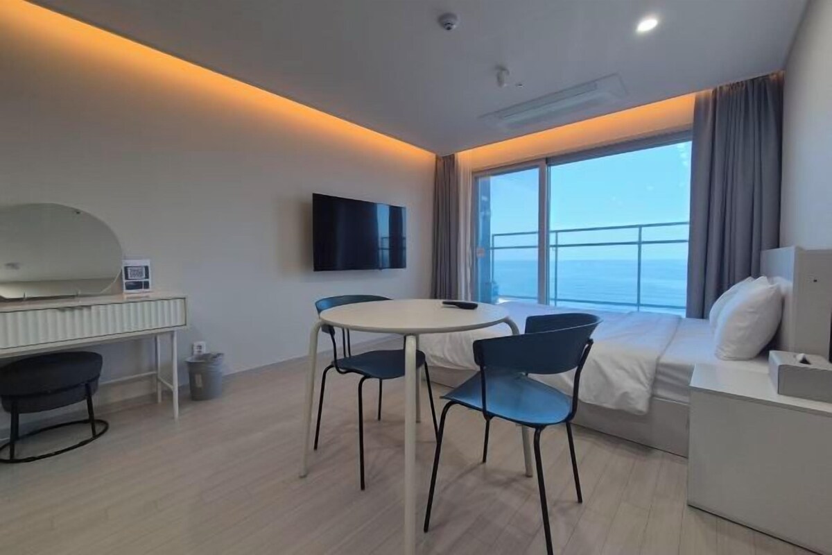 A modern hotel room with a bed, a desk, a chair, a television, and a large window offering a view.