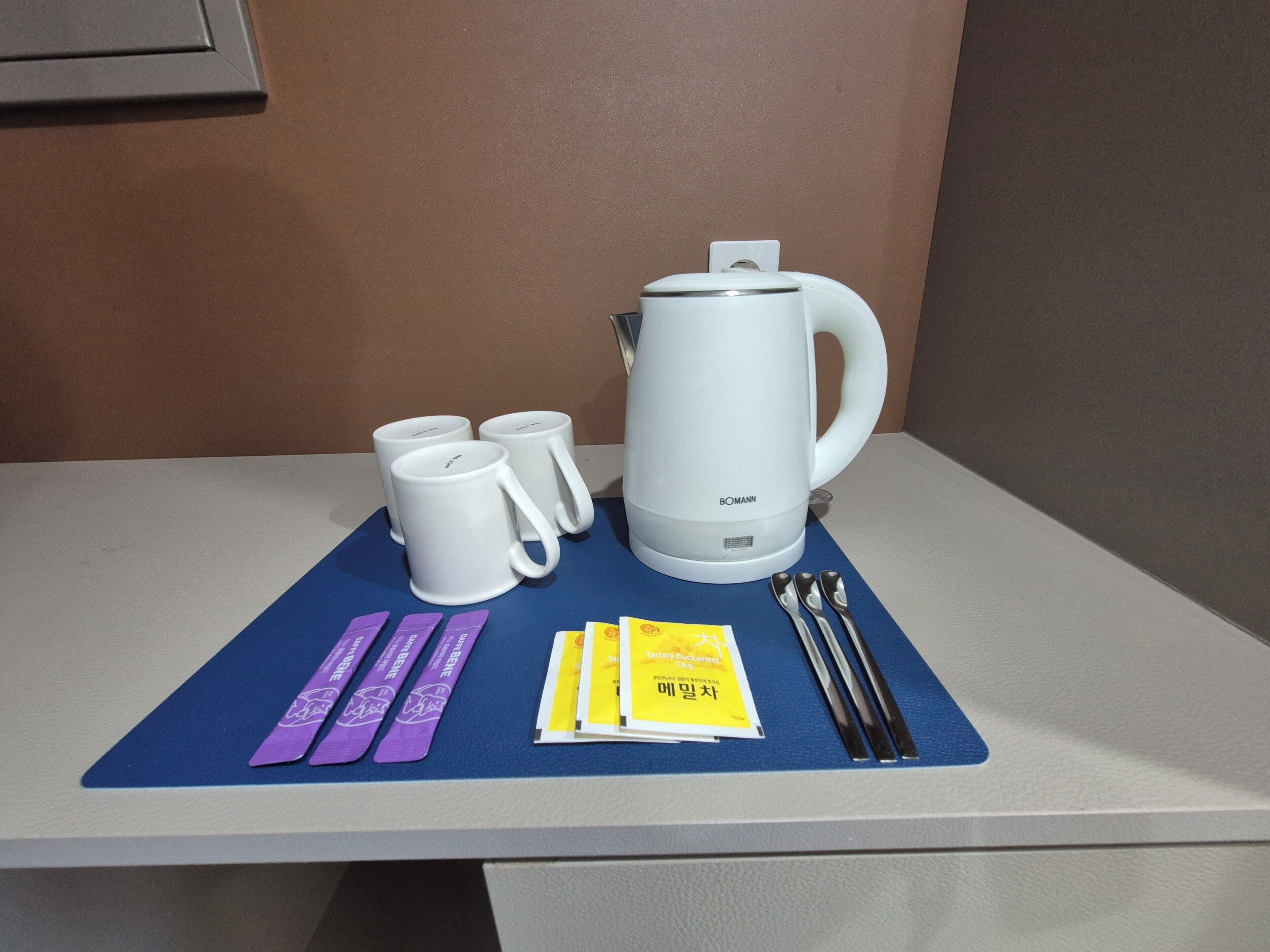 Une table sur laquelle se trouvent une bouilloire électrique blanche, trois tasses blanches, un set de table bleu, un petit livret jaune et deux bracelets violets.