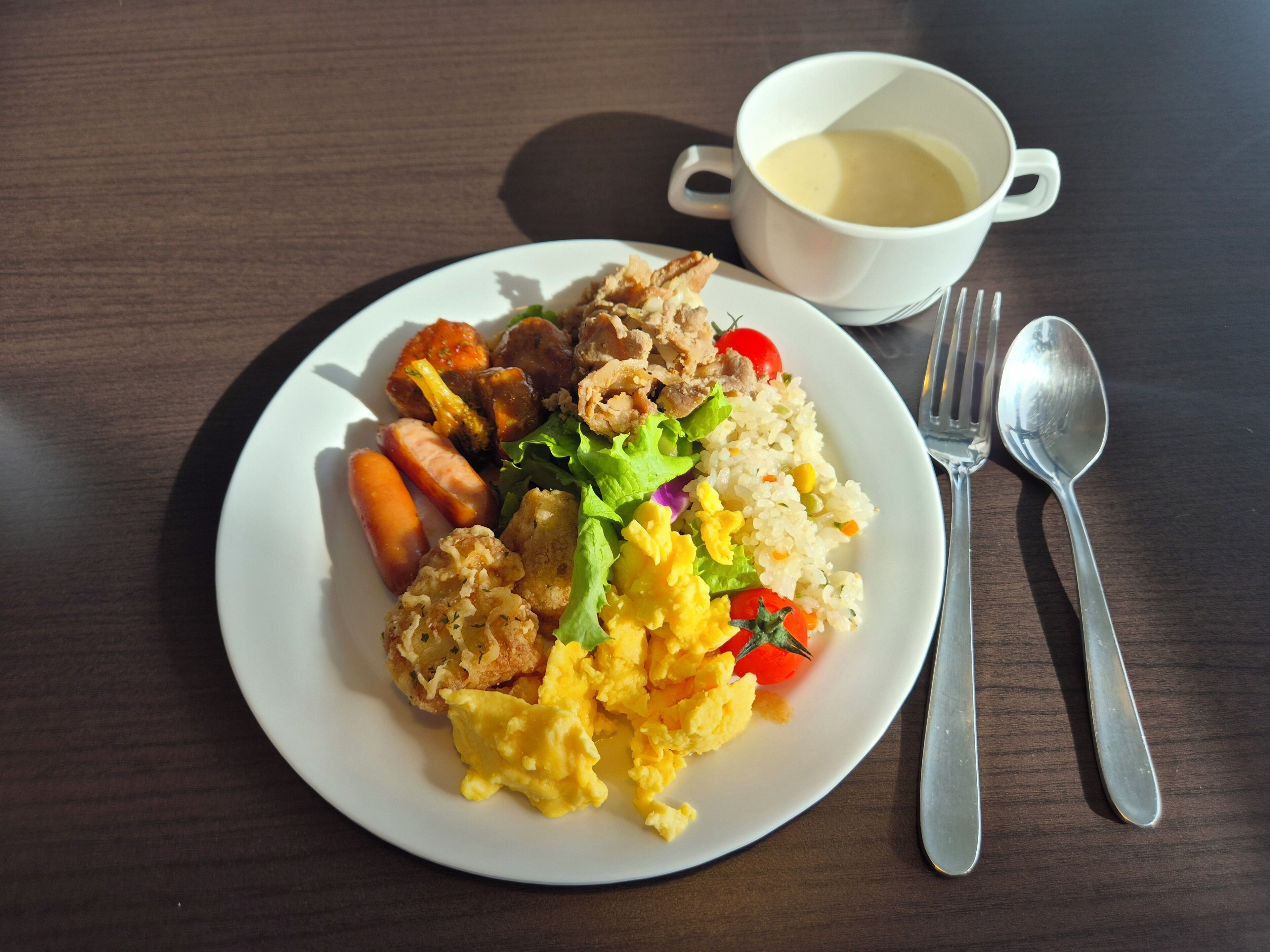 A plate of food with scrambled eggs, sausage, and vegetables, accompanied by a cup of soup and cutlery on a wooden table.
