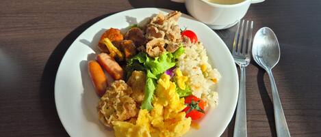 A plate of food with scrambled eggs, sausage, and vegetables, accompanied by a cup of soup and cutlery on a wooden table.