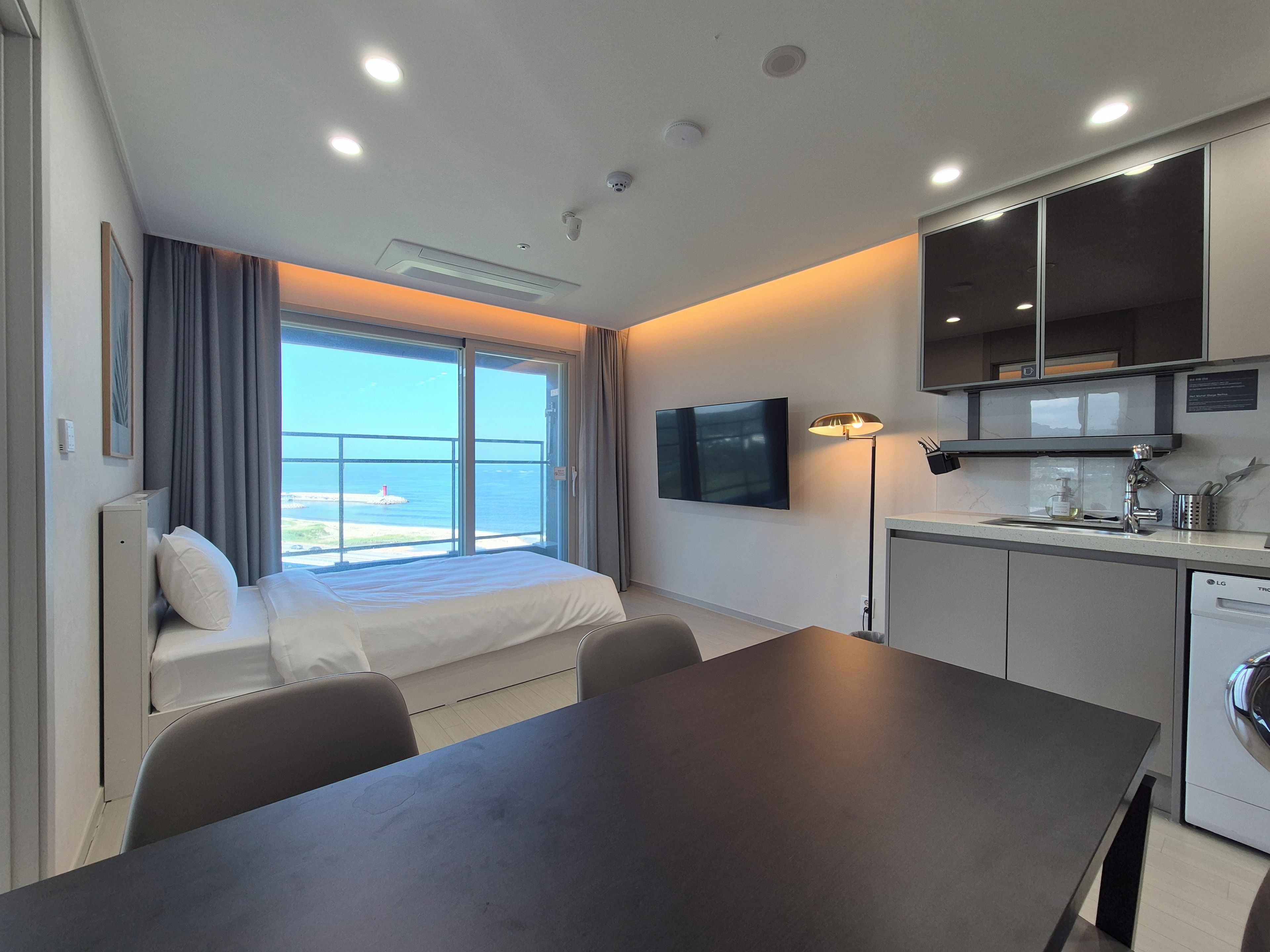 A modern hotel room with a bed, a dining table, a kitchen area, and a large window offering a view of the sea.