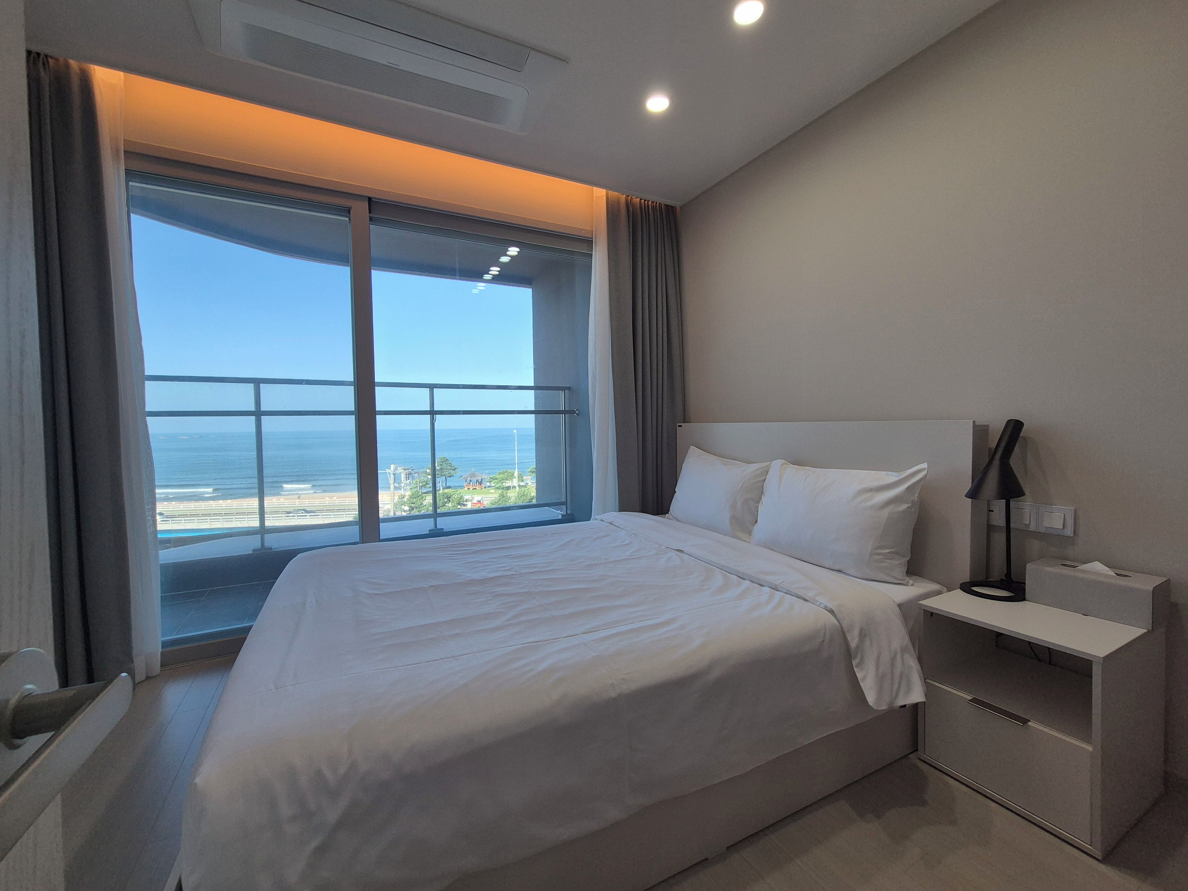 A hotel room with a large bed, a bedside table, a lamp, and a view of the ocean.