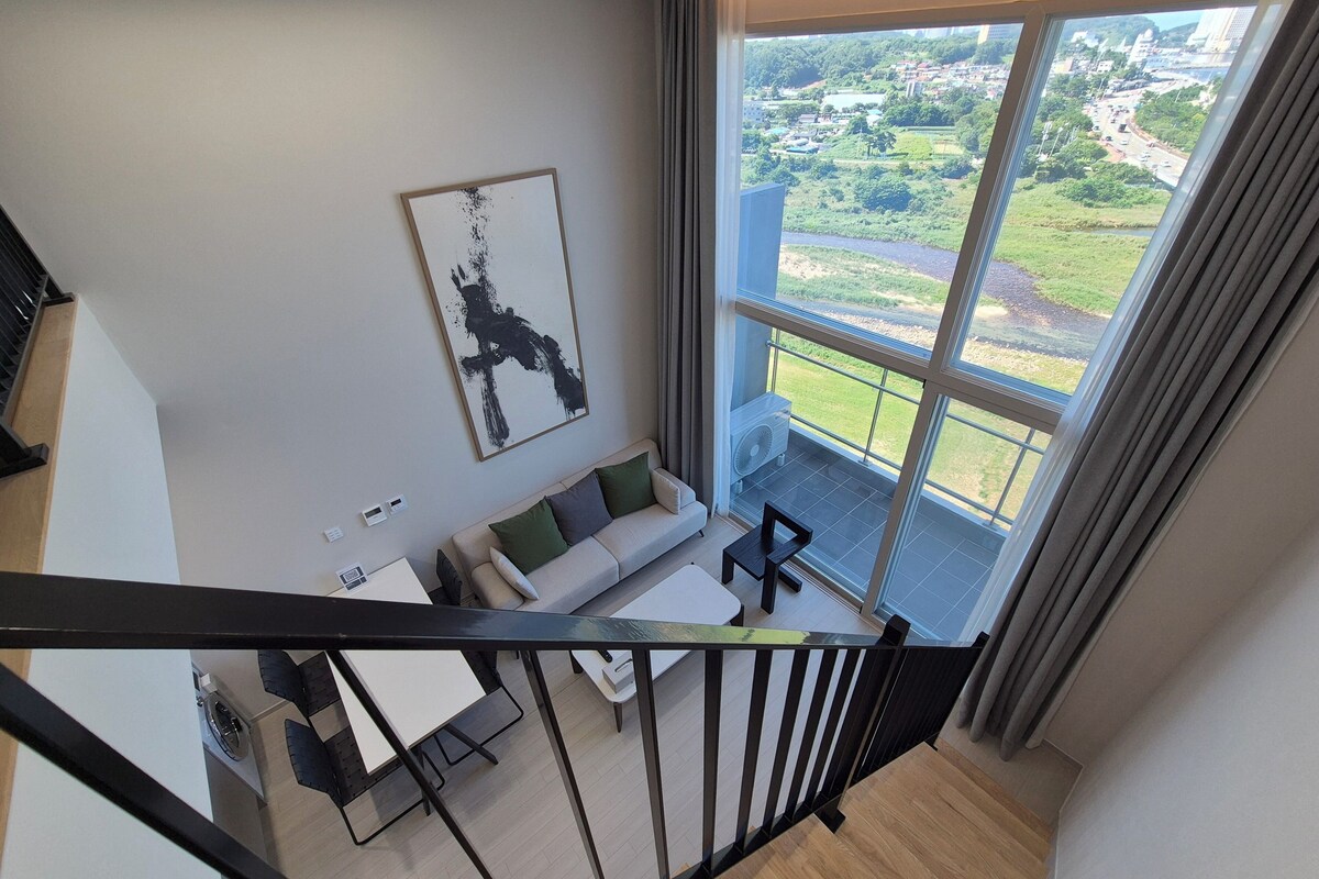 A modern living room with a sofa, a coffee table, and a view of a green landscape through large windows.