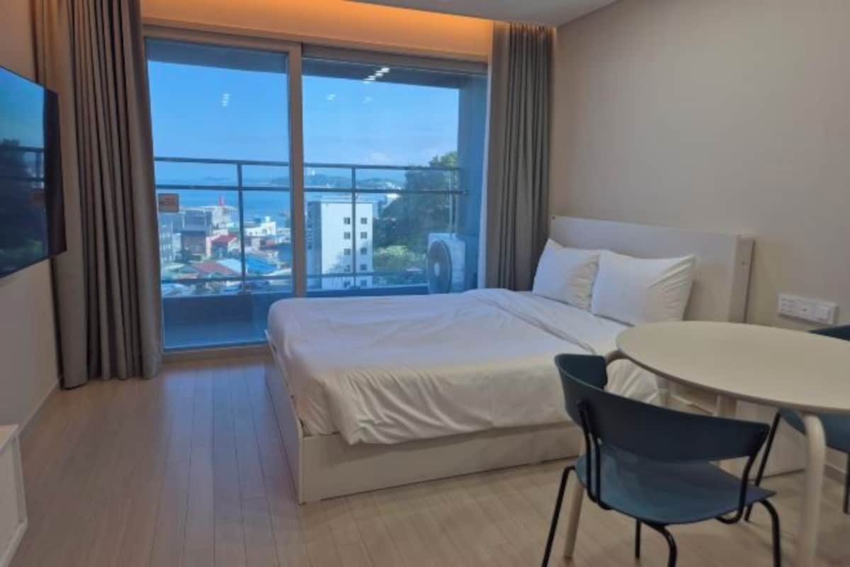 A hotel room with a bed, a table, and a chair. There is a balcony with a view of the city.