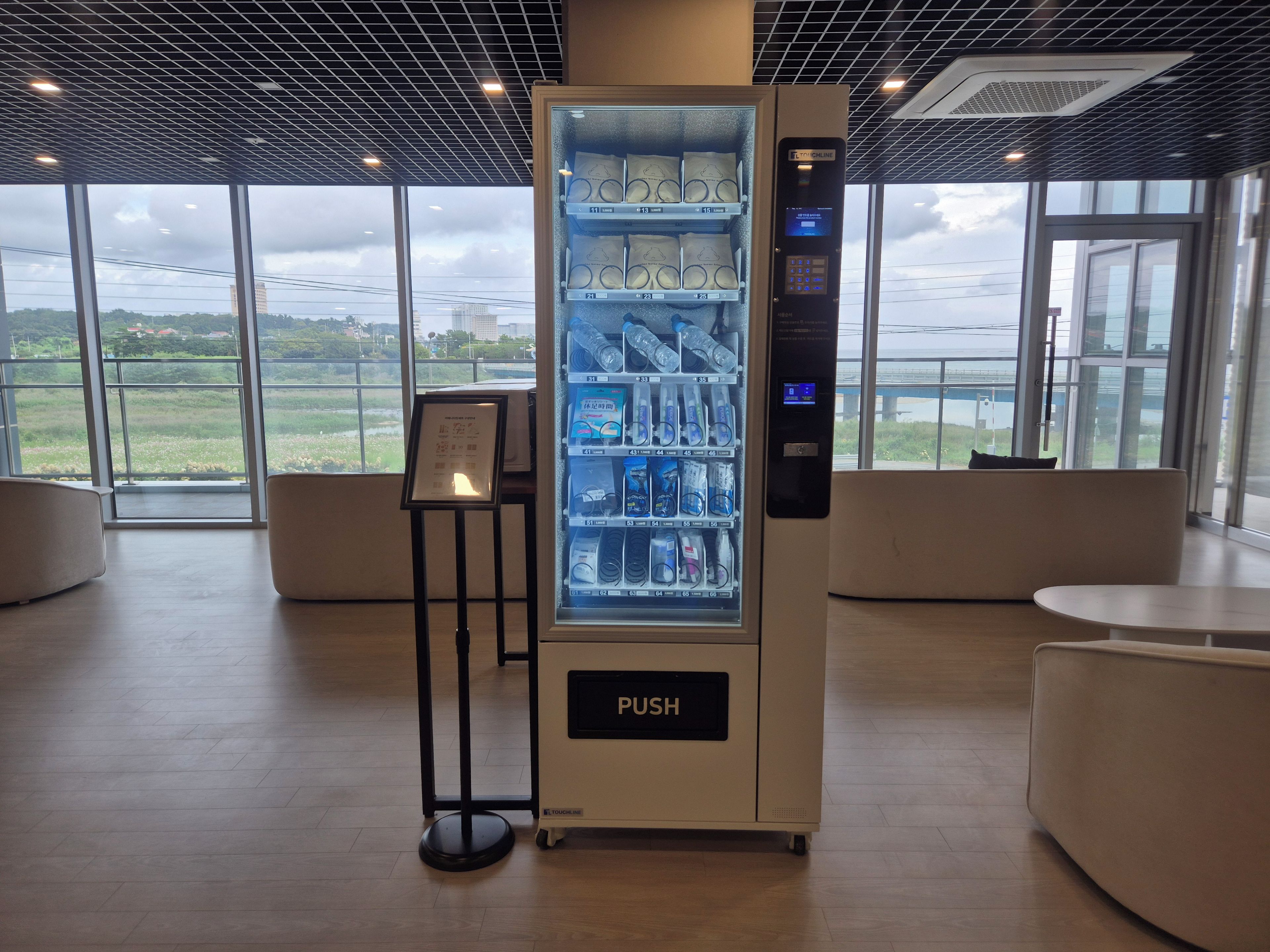 A vending machine with a push button, located in a room with large windows and a view of a distant landscape.