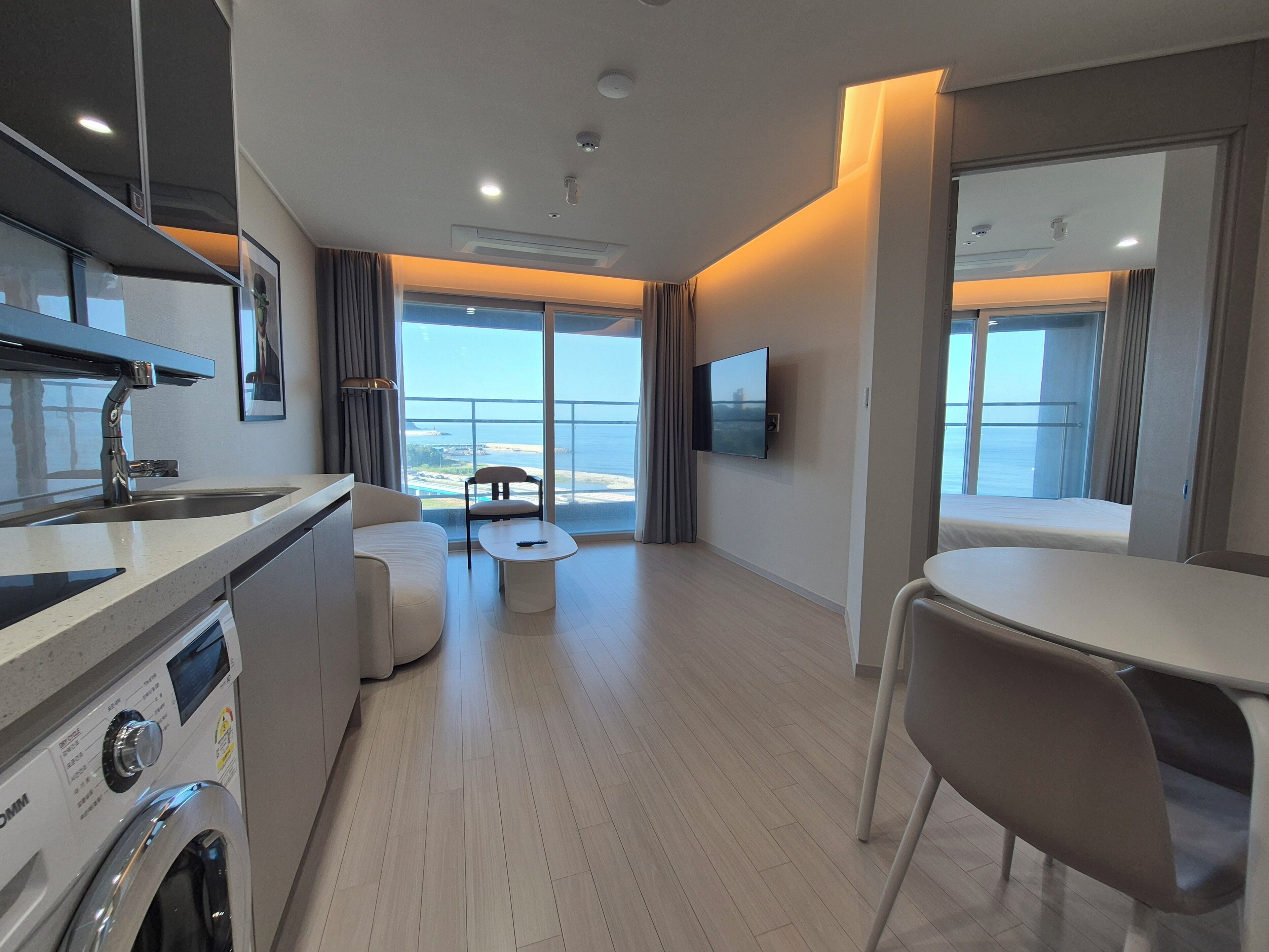 A modern hotel room with a kitchen area, a dining table, a sofa, and a large window offering a view of the sea.