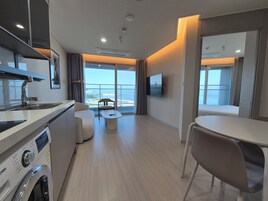 Habitación de hotel moderna con cocina, una mesa de comedor, un sofá y un amplio ventanal con vistas al mar.