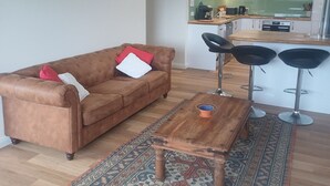 A living room with a brown sofa, a coffee table, and a patterned rug. There is a painting on the wall and a kitchen area in the background.