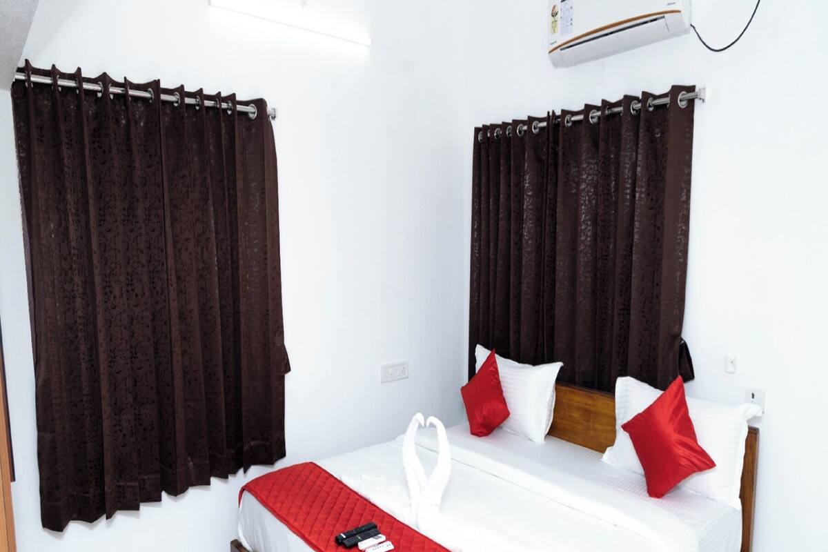 A hotel room with a bed, curtains, a red blanket, pillows, and a remote control on the bed.
