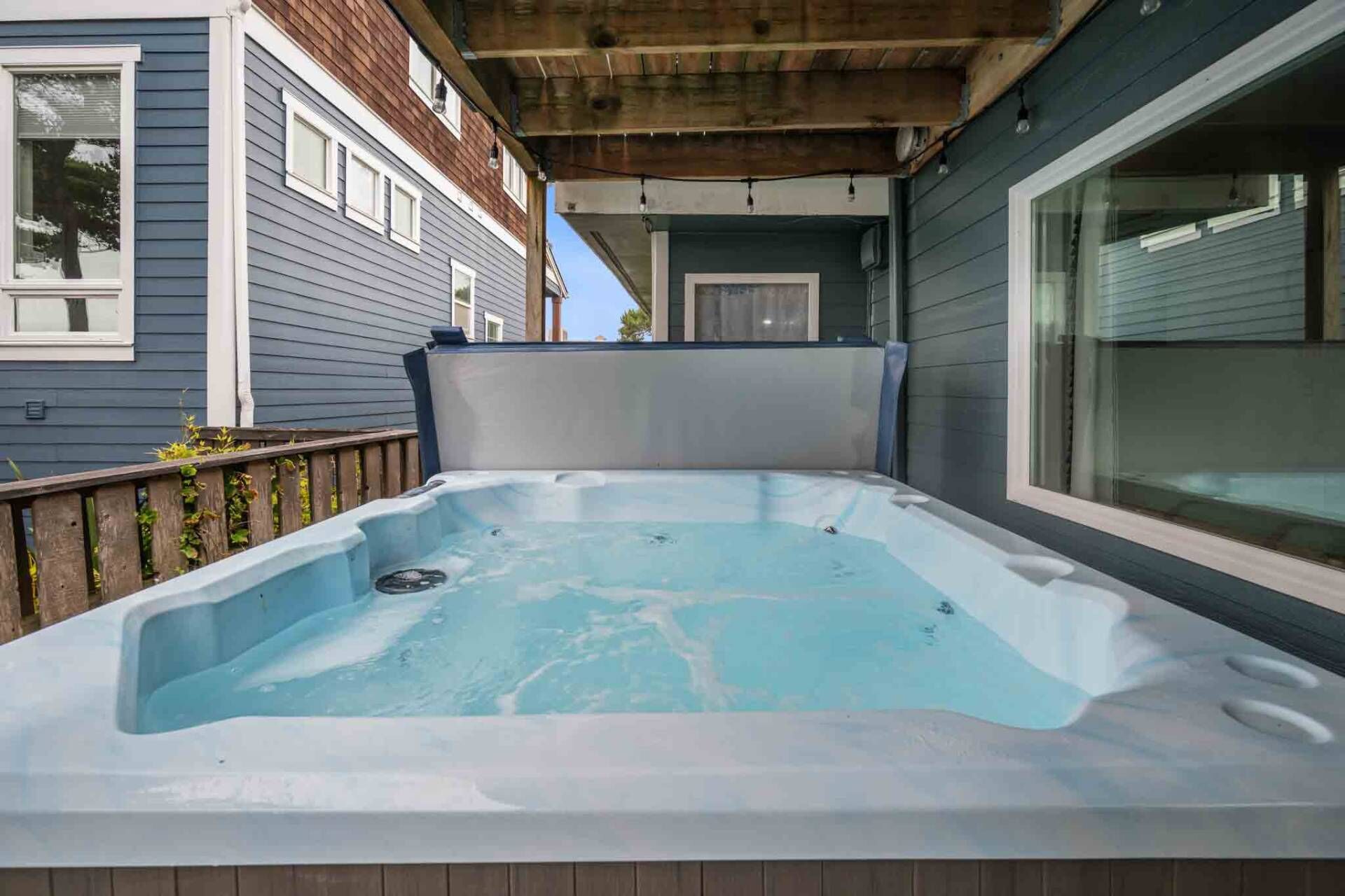 A hot tub in a covered outdoor area with wooden decking and a view of a house with blue siding and a window.