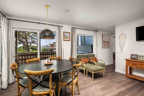 A dining area with a round table, wooden chairs, and a pendant light. There is a window with curtains and a view of greenery outside.