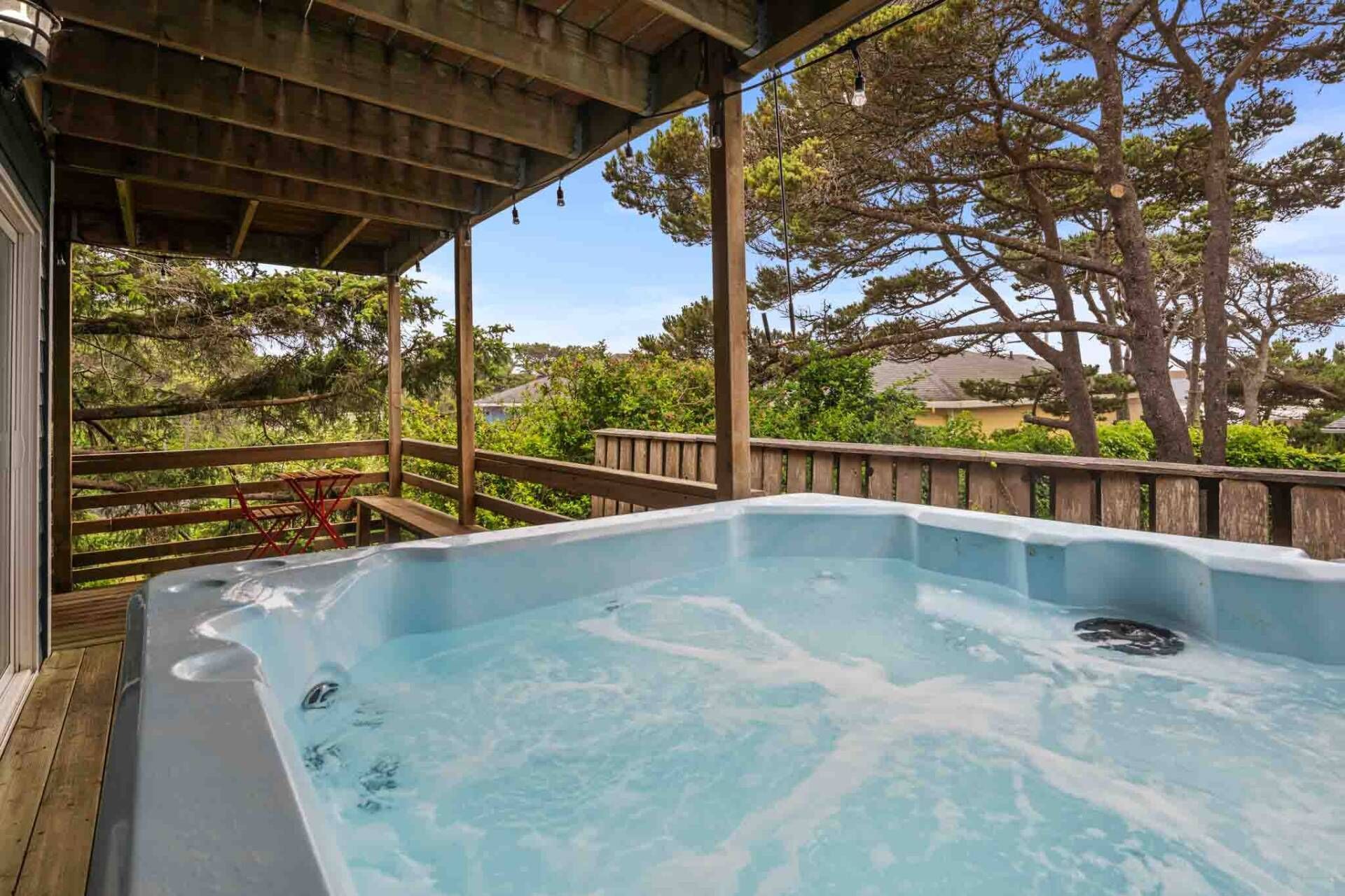 Un jacuzzi debajo de una terraza de madera, con vistas a árboles y una casa al fondo.