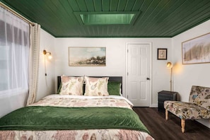A bedroom with a green upholstered bed, a side table, a chair with floral upholstery, a painting on the wall, and a door.